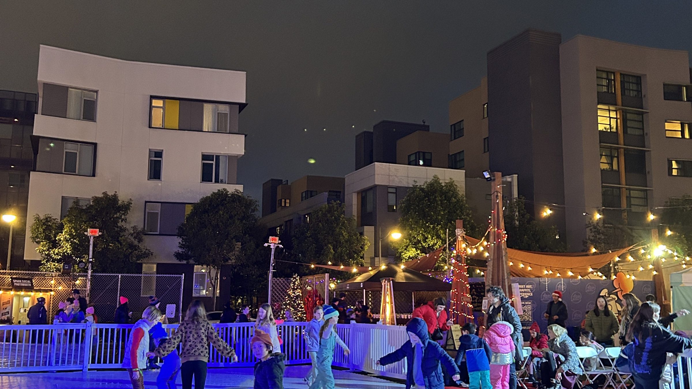 Children ice skating at night under blue lights, with buildings in the background.