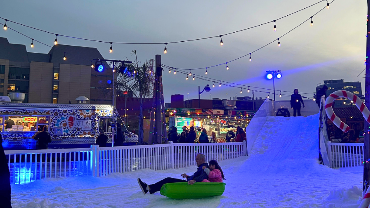 Adults and children sledding on artificial snow slide, surrounded by lights and city buildings at dusk.