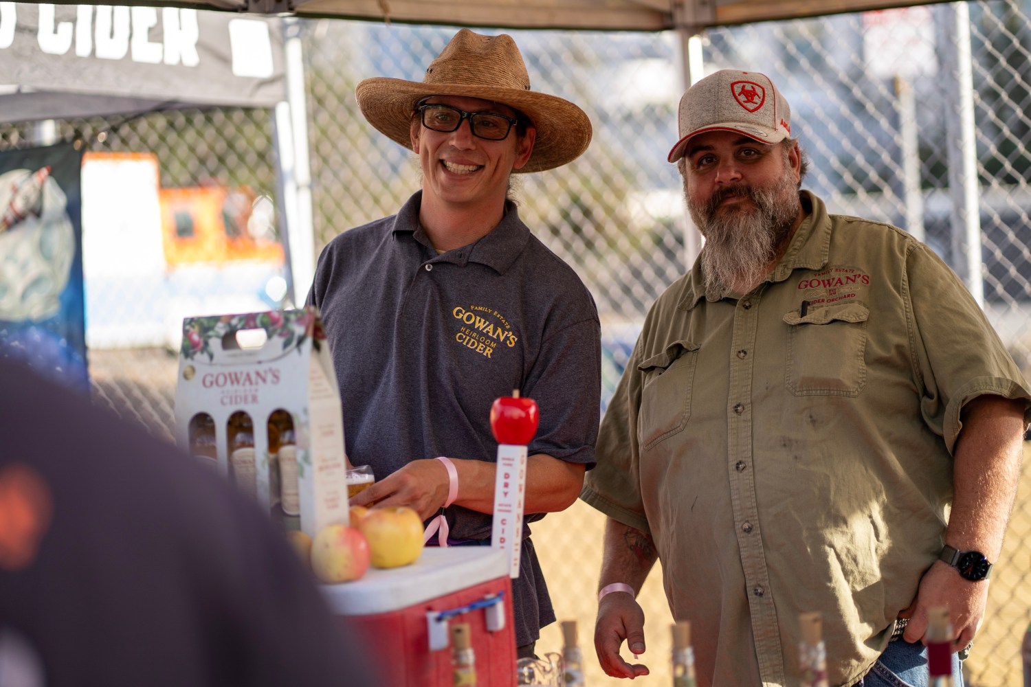 Two men in hats at a cider booth with apples and bottles displayed.