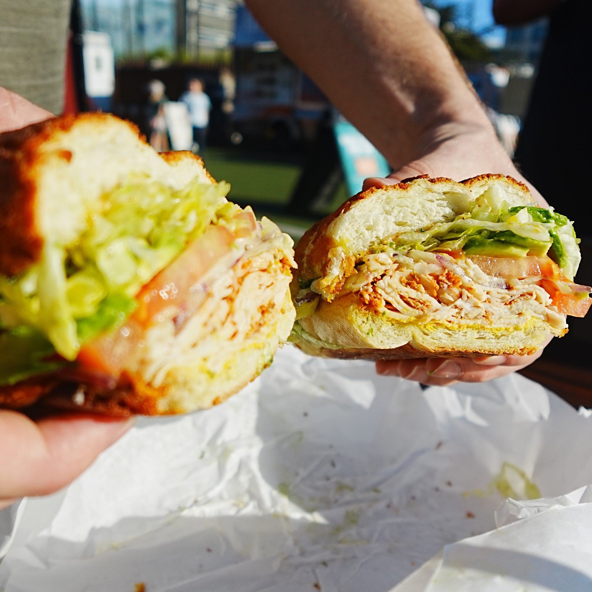 Person holding a cut sandwich with lettuce and tomato, outside on a sunny day.
