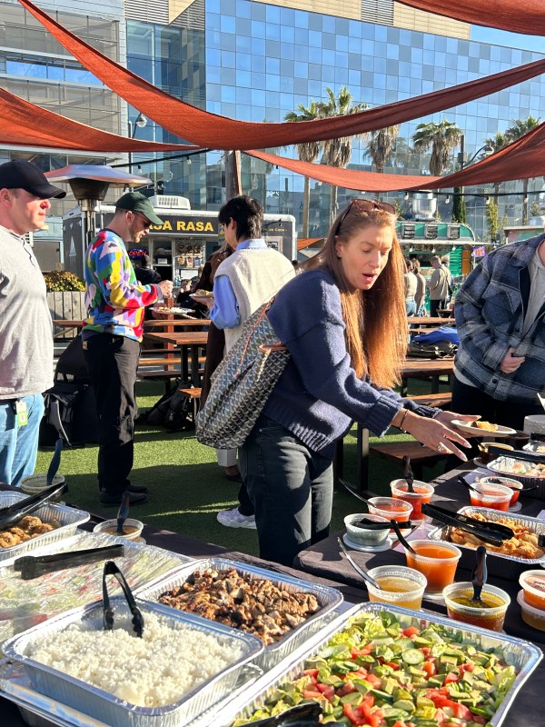 People serving food from a buffet table outdoors with buildings in background.